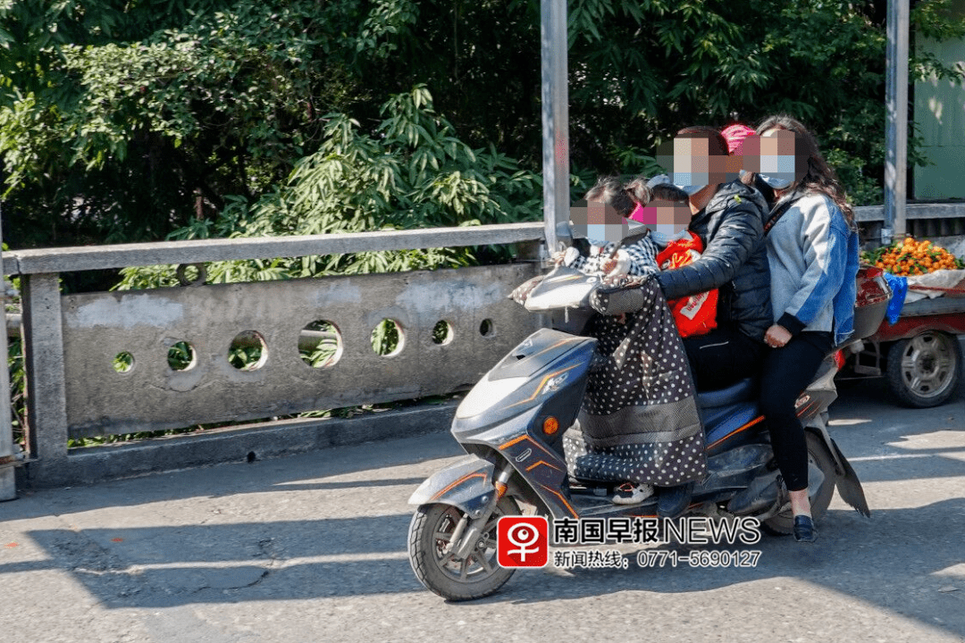 一名女子骑着电动车搭载两个孩子,一个站在踏板上,另一个坐在后座